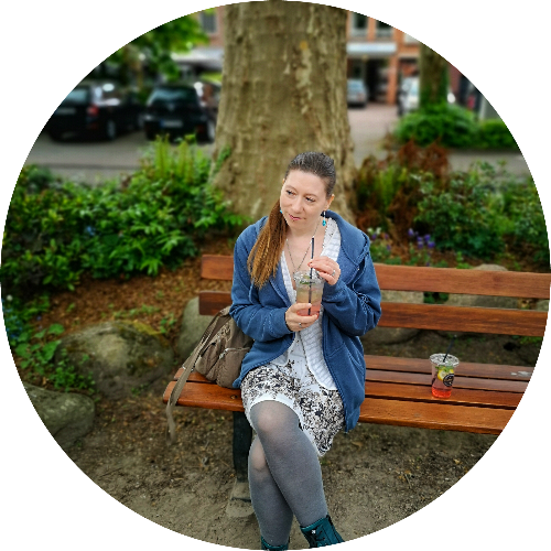 Bild von mir. Ich sitze auf einer Bank draußen und halten einen To-Go-Becher in der Hand. Ich trage ein geblümtes Kleid und eine blaue Strickjacke. Meine braunen, langen Haare sind zum Pferdeschwanz gebunden.